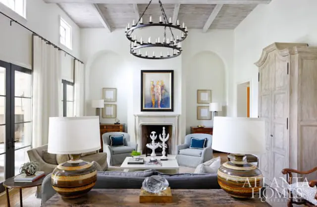 Living room with white walls, beamed ceiling, and a large fireplace. Features include two circular chandeliers, upholstered armchairs, a white coffee table, and decorative lamps.