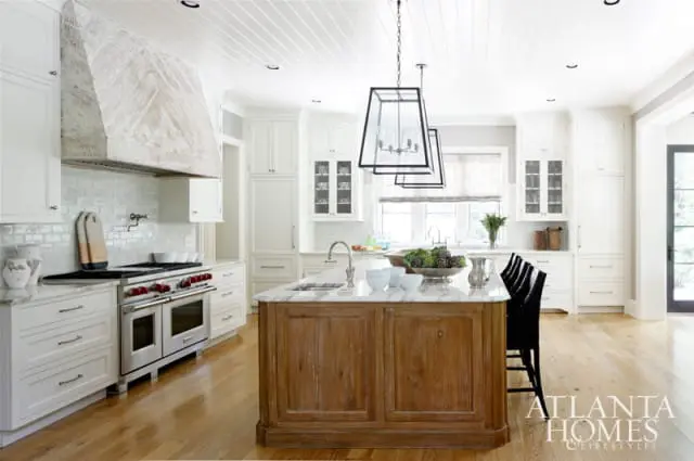 White kitchen with a large stone range hood, marble island with a wooden base, and pendant lights.