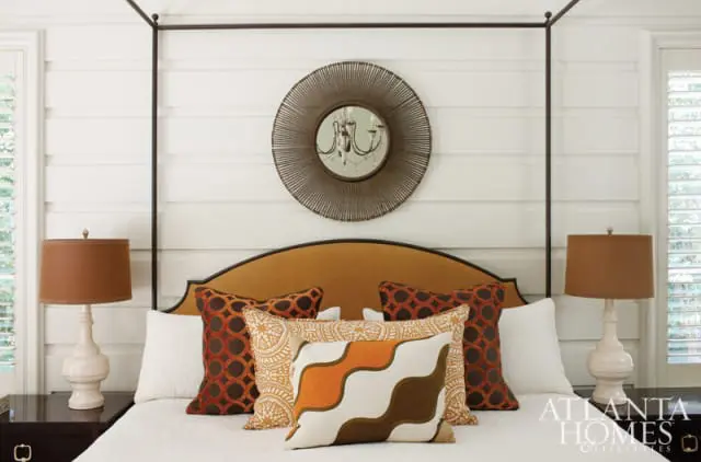 Bedroom with a four-poster bed, upholstered headboard in rust orange, and white shiplap walls. Pillows feature geometric and abstract patterns in rust orange, brown, and cream. A round metal sunburst mirror hangs above the bed.