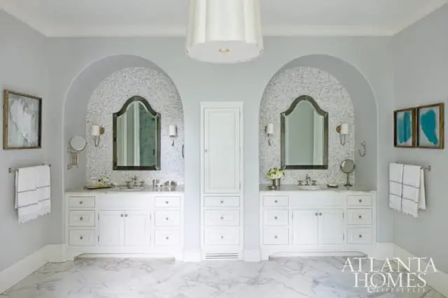 Light gray bathroom with twin white vanities, arched niches with mosaic tile backsplashes, ornate mirrors, marble floors, and a tall white linen cabinet