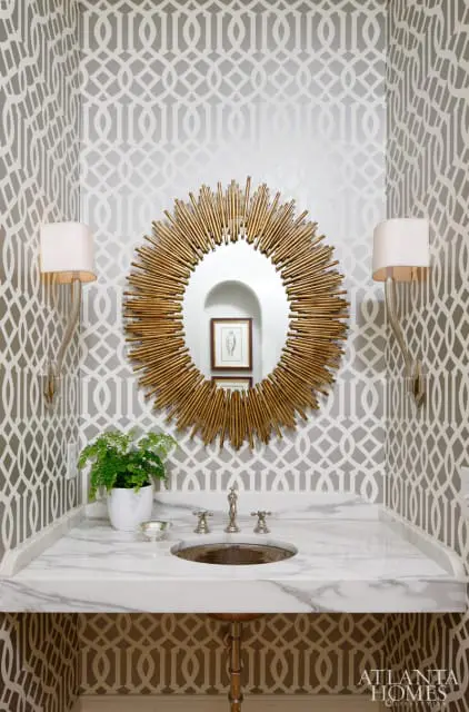 Powder room with gray and white geometric wallpaper, a marble vanity with a bronze sink, a sunburst mirror, and two sconces.