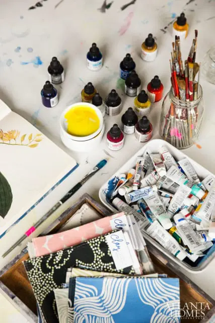 Various art supplies including ink bottles, paint tubes, brushes, fabric swatches, and an open sketchbook with floral illustrations.
