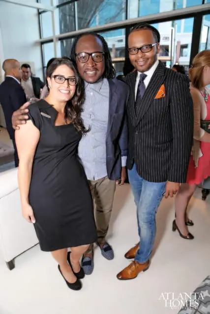 Three people posing for a photo indoors. A woman in a black dress smiles next to two men, one in a pinstripe suit and the other in a blue striped shirt and blazer.