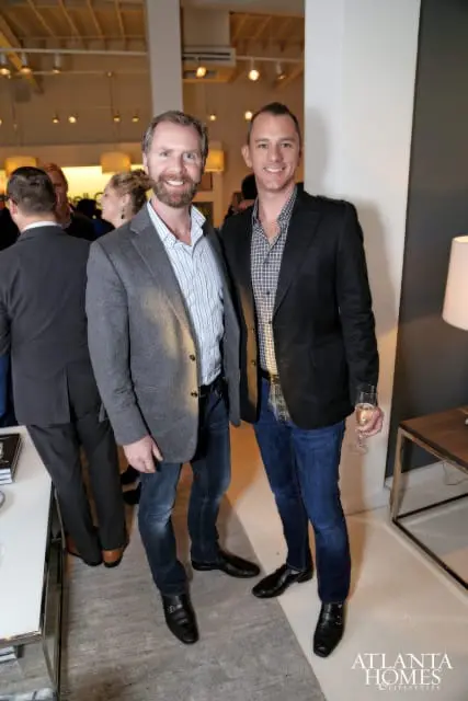 Two men in blazers and jeans smile at the camera. The man on the left wears a gray blazer over a striped shirt. The man on the right wears a black blazer over a plaid shirt and holds a champagne flute.