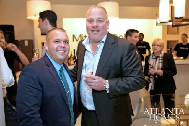 Two men in suits holding champagne flutes at a social gathering, with other attendees and a bar in the background.