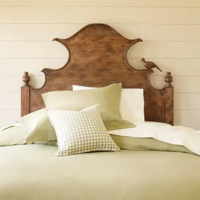 Bedroom with a wooden headboard featuring carved details and a bird accent, dressed in light green bedding and a gingham pillow.