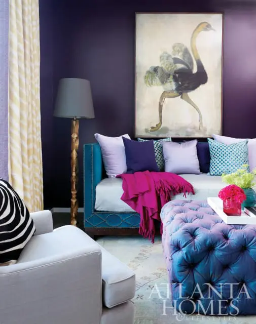 Living room with deep purple walls, teal daybed with jewel-toned pillows and a fuchsia throw, and a tufted ottoman.