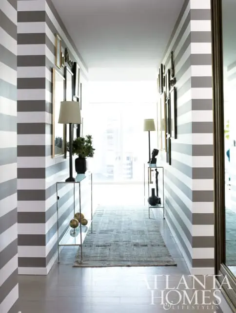 Hallway with gray and white striped walls, a mirrored wall, framed art, console tables with lamps and sculptures, and a patterned runner rug on hardwood floors.