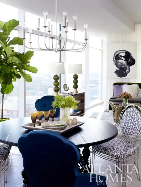 Round black dining table with navy velvet chairs and patterned upholstered chairs, set with a tray of amber glasses and green floral centerpiece.