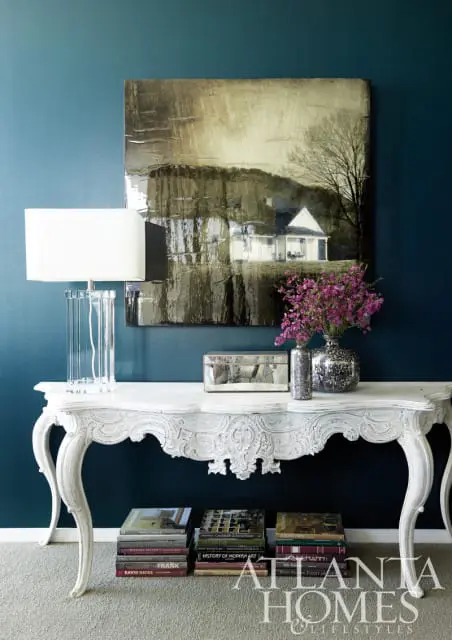White ornate console table with a glass lamp, mirrored box, silver vase with purple flowers, and stacked books on a teal wall.
