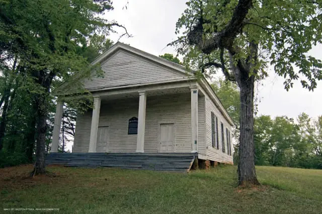 Weathered white clapboard church with a pedimented gable, Doric columns, and double doors, surrounded by trees and grass.