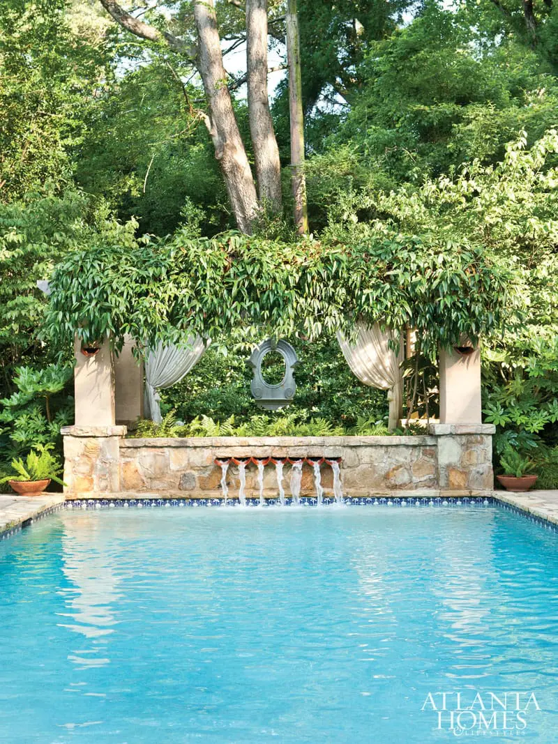 Blue swimming pool with a stone wall featuring a row of seven water spouts cascading into the pool, surrounded by lush green trees and foliage.