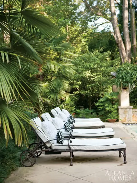 Row of white chaise lounges with black trim and decorative pillows beside a pool, surrounded by lush palm trees and greenery.