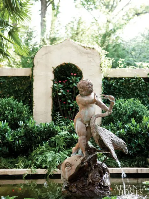Bronze fountain sculpture of a cherub with a bird in a garden pond, with lush greenery and an arched wall in the background.
