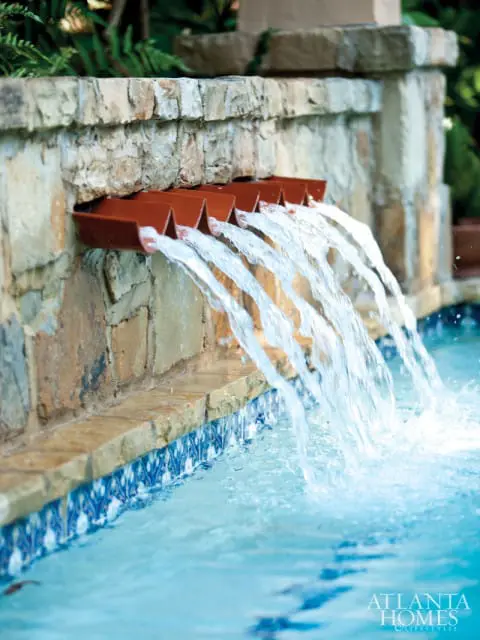Stone wall with multiple copper spouts cascading water into a blue tiled swimming pool with lush greenery in the background.