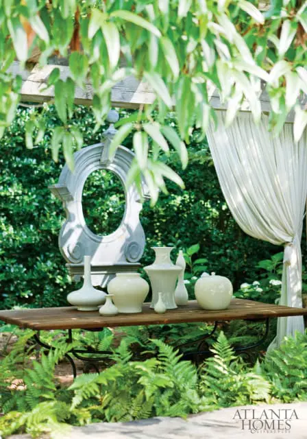 Garden setting with a wooden table displaying cream-colored vases and a stone architectural element, surrounded by lush greenery and ferns.