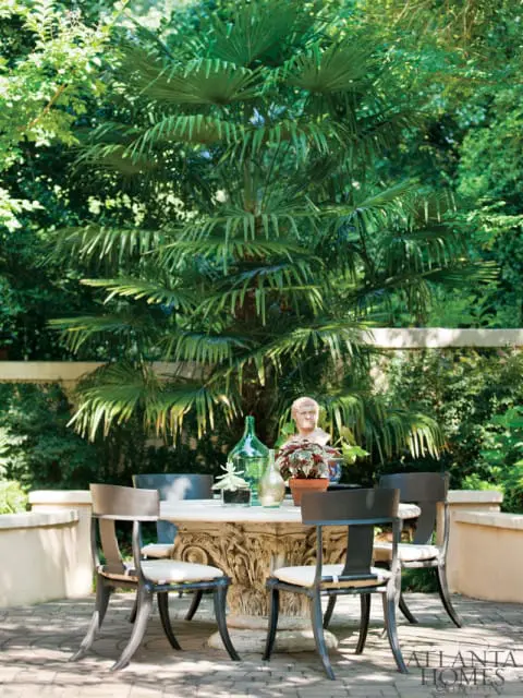 Stone patio with a round table and four chairs, decorated with glass bottles, plants, and a bust, set against a backdrop of lush greenery and a palm tree.