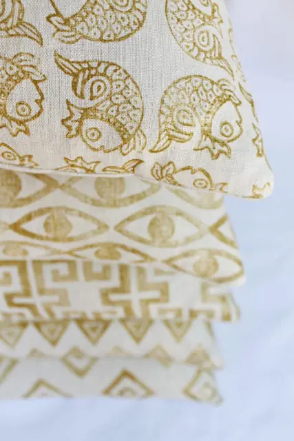 Stack of four pillows with gold patterns on a white background. Top pillow features a fish pattern, second has an eye pattern, third has a Greek key pattern, and the bottom has a geometric triangle pattern.