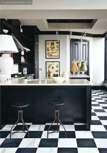 Black kitchen island with marble countertop, two industrial stools, and a checkered black and white marble floor. Vintage botanical prints hang on the wall.