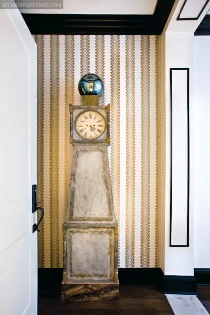 Entryway with a tall, distressed grandfather clock against a wall with gold leaf fern-patterned wallpaper and black trim