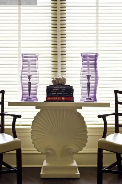 Console table with a shell-shaped pedestal, topped with books and two purple glass hurricane candle holders, flanked by upholstered chairs.