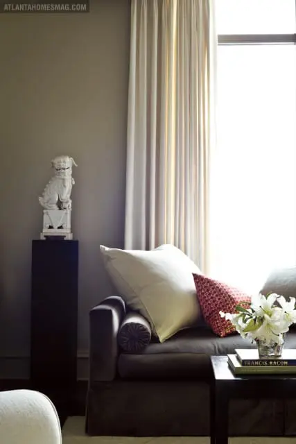 Dark gray sofa with cream and patterned red pillows, a white foo dog statue on a pedestal, and a vase of white lilies on a side table with books.