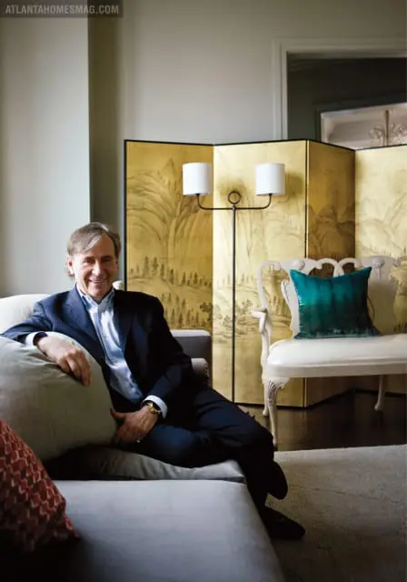 Man in a dark pinstripe suit and light blue dress shirt sitting on a grey sofa, with a gold folding screen and a white bench with a teal pillow in the background.