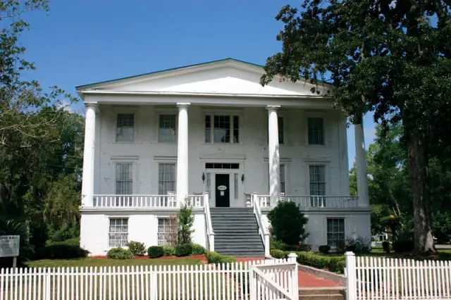 White neoclassical mansion with a portico supported by large columns, a wide staircase leading to the entrance, and a white picket fence in the foreground.
