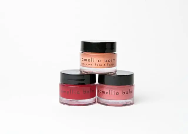 Three small glass jars of camellia balm with black lids. One jar is peach colored, one is red, and one is pink.