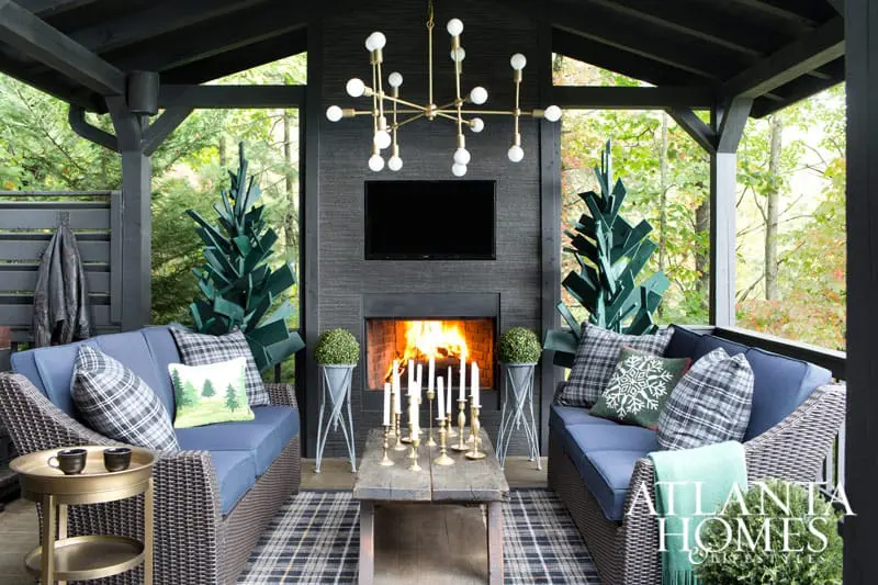 Covered porch with blue upholstered sofas, plaid pillows, a fireplace with a TV above, and decorative green trees.