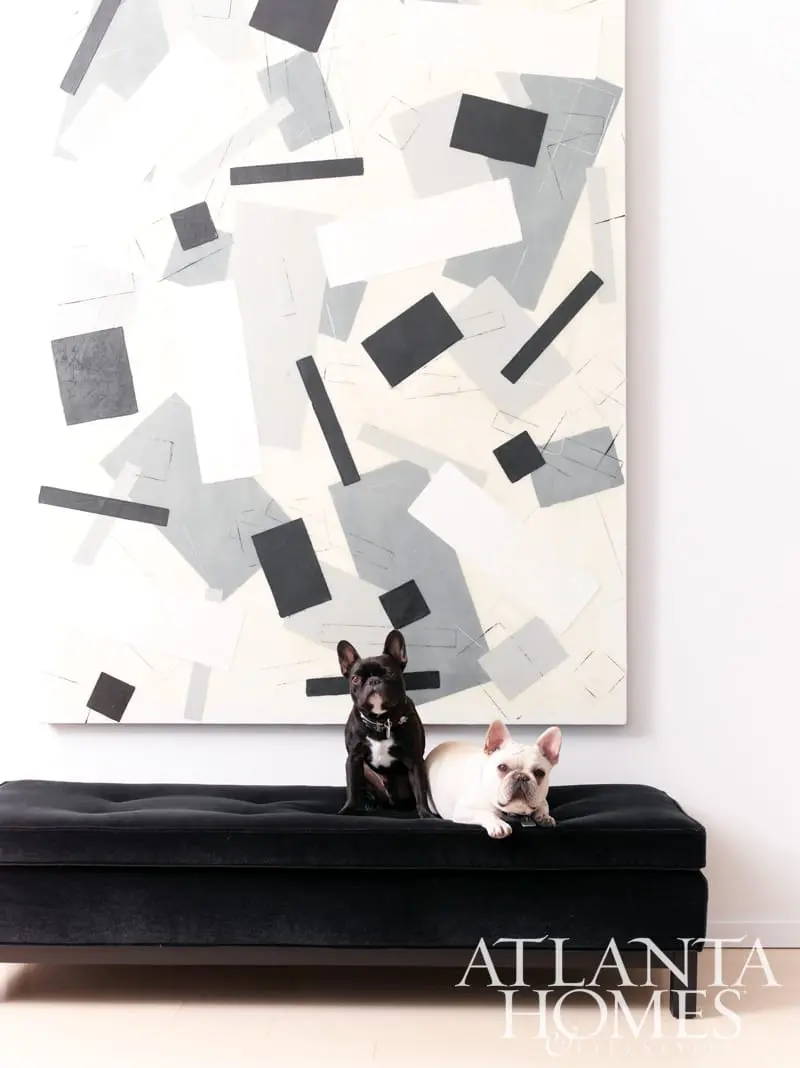 Two French bulldogs sit on a black tufted bench in front of a large abstract painting with geometric shapes in black, white, and gray.