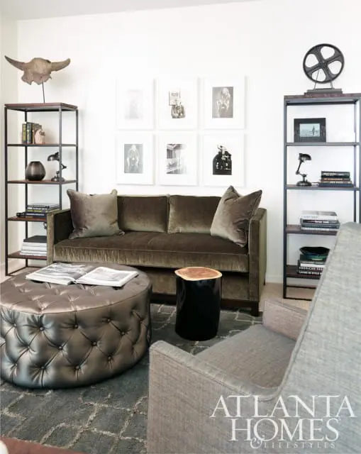 Living room with a plush olive green sofa, tufted metallic ottoman, and grey armchair. Wall features a gallery of black and white photographs and decorative shelving.