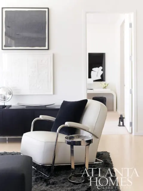 Living room with a white leather armchair, black velvet pillow, and chrome side table on a gray rug. A black credenza with artwork hangs on the wall.