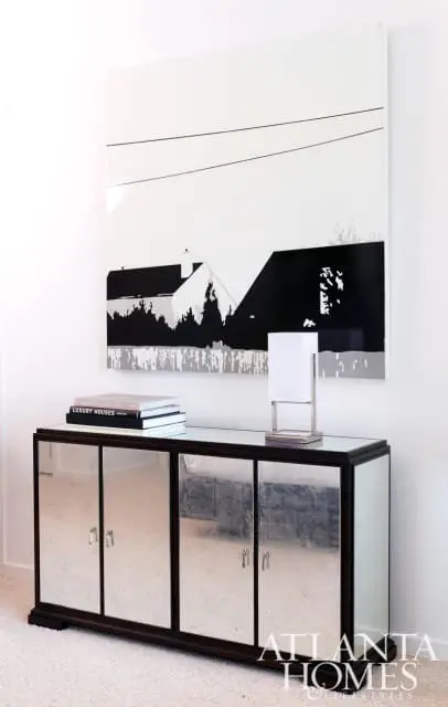 Modern living room with mirrored cabinet, stack of books, and abstract art featuring houses and power lines.