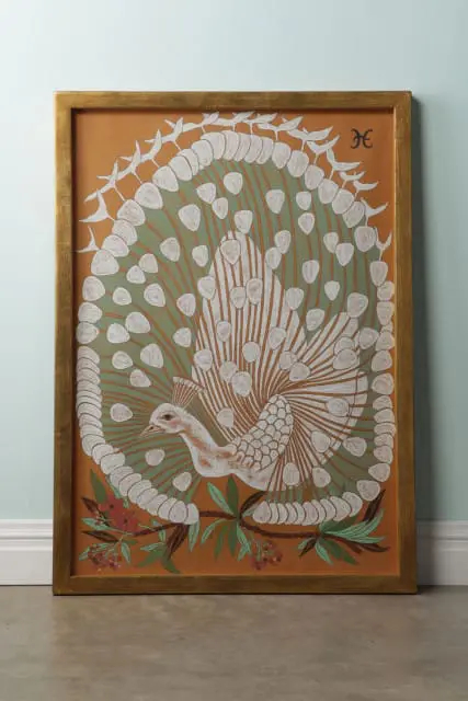 Framed artwork of a peacock with its tail feathers fanned out, rendered in white, green, and orange hues on an orange background. A monogram 'H' is visible in the upper right corner.