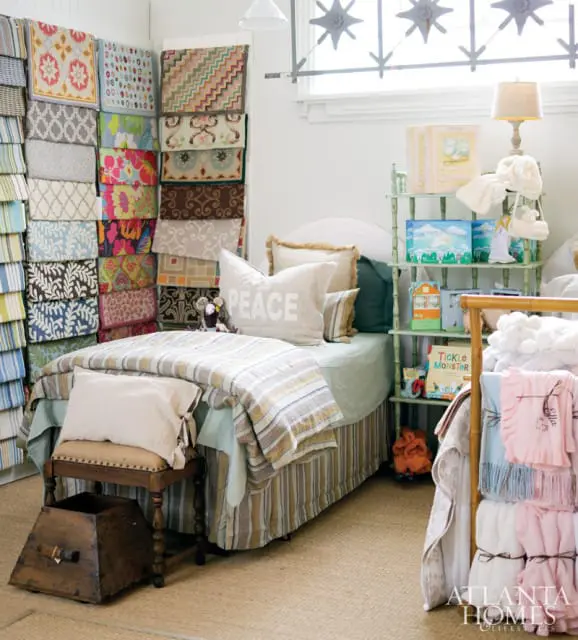 Bedroom with a twin bed, stacks of fabric samples, and shelves of children's books and toys. The bed is covered in striped bedding and a pillow that says 'PEACE'.