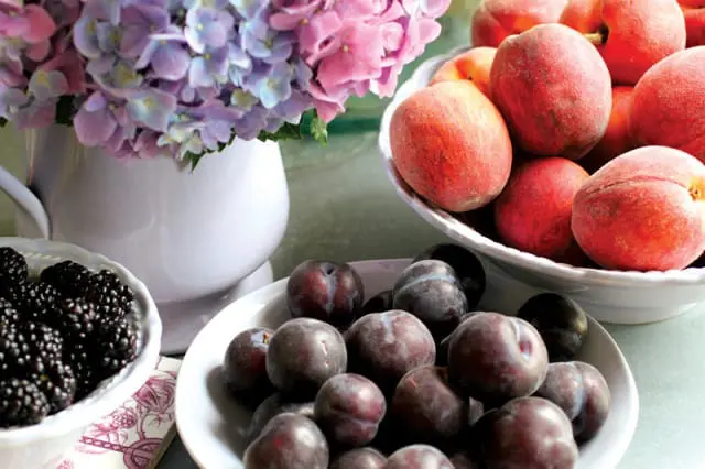 Bowls of fresh blackberries, plums, and peaches arranged with a vase of pink and purple hydrangeas.