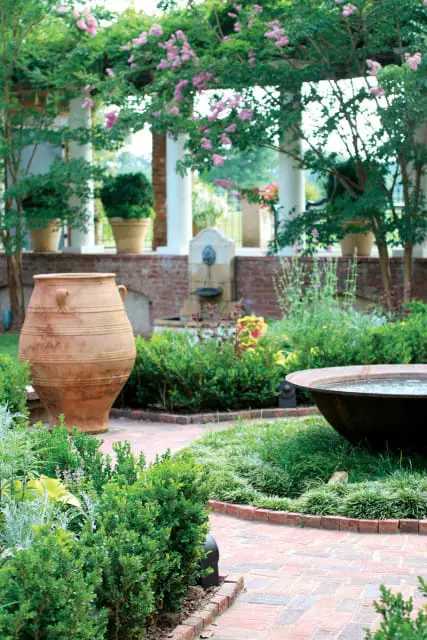 Terracotta urn and weathered metal basin in a garden with brick pathways, manicured hedges, and flowering vines on a pergola.