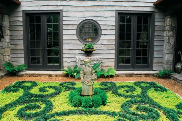 Stone statue of a woman holding a basket in a garden with swirling green and yellow plants, flanked by French doors and a round window on a wood-shingled wall.