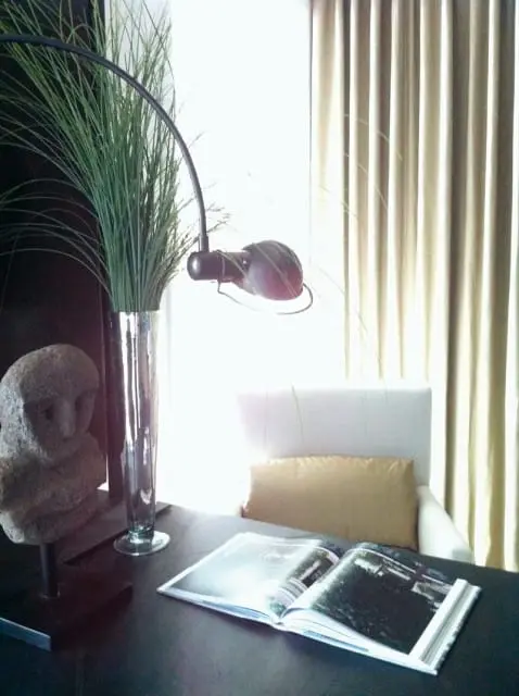 Desk with an open book, a stone bust, a tall glass vase with green grass, and a modern lamp. A white armchair with a tan pillow sits behind the desk, with light-colored curtains in the background.