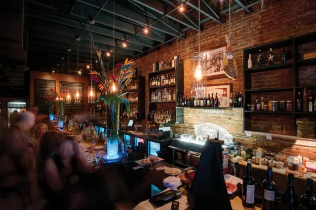 Bar interior with brick walls, shelves stocked with bottles, and a long bar with patrons. A large vase of tropical flowers sits on the bar.