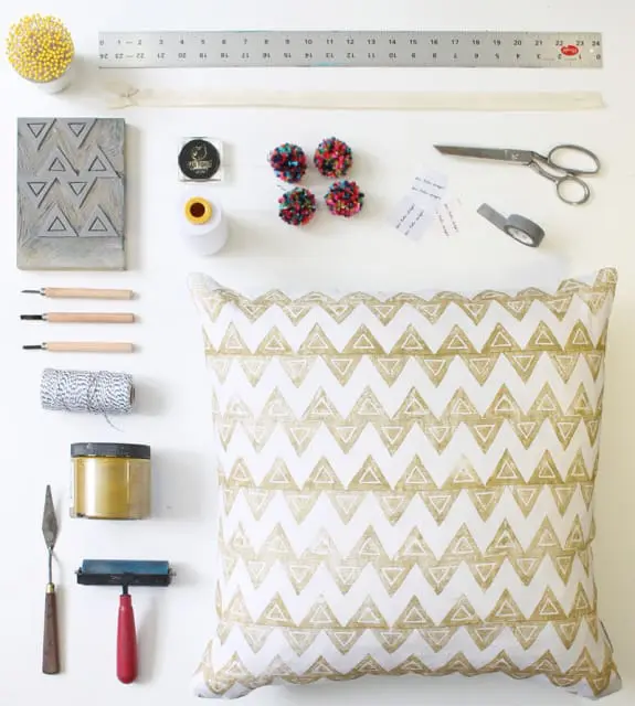 White pillow with a repeating pattern of gold triangles, surrounded by craft supplies including a ruler, carving tools, twine, gold paint, a roller, scissors, tape, and pom-poms.