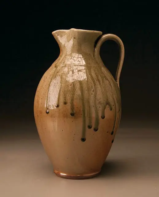 Earthenware pitcher with a light brown glaze and green drips, featuring a curved handle and a wide spout.