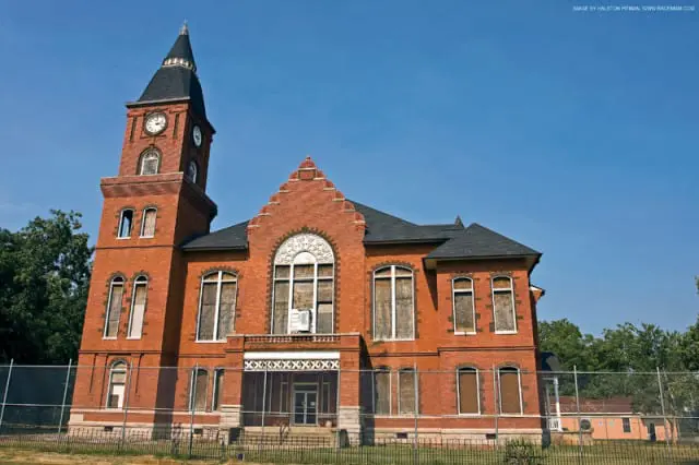 Red brick courthouse with a clock tower, arched windows, and boarded-up windows under a clear blue sky.
