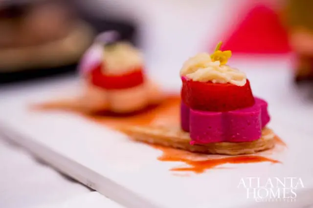 Close-up of a small appetizer with layers of pink, red, and a creamy topping, served on a cracker with red sauce and a white rectangular plate. Atlanta Homes & Lifestyles logo in the bottom right.