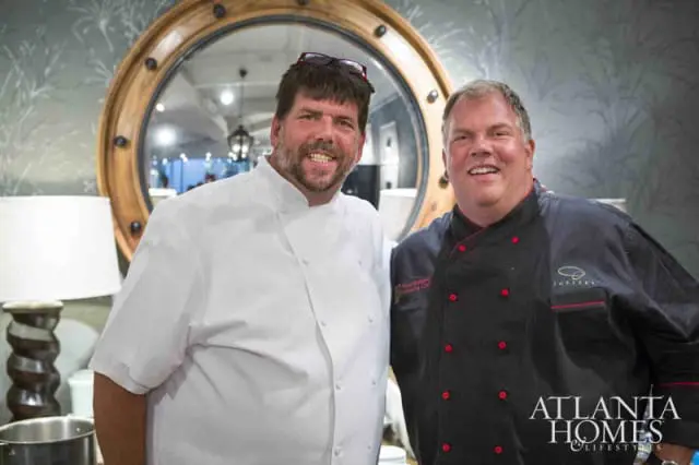 Two chefs in chef coats smile in front of a large round mirror with a wooden frame. The chef on the left wears a white coat, and the chef on the right wears a black coat with red trim.