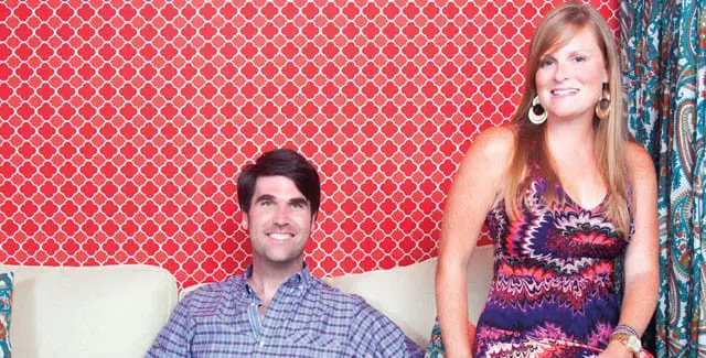 Man in blue plaid shirt and woman in colorful patterned dress smile in front of red quatrefoil wallpaper and patterned curtains.