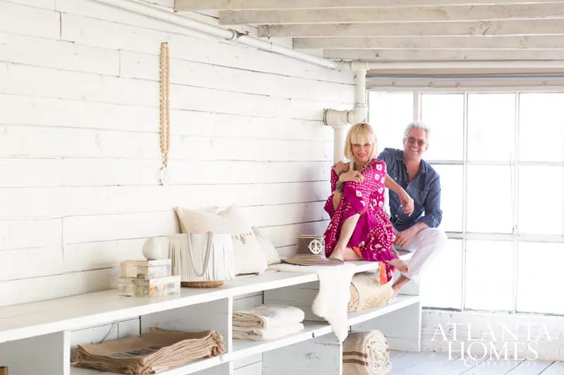 A couple sits on a white bench in a room with white shiplap walls and large industrial windows. The woman wears a pink patterned dress and the man wears a denim shirt and white pants.
