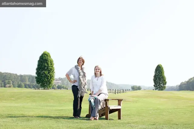 Two women stand and sit on a wooden chair in a grassy field with trees and rolling hills in the background.