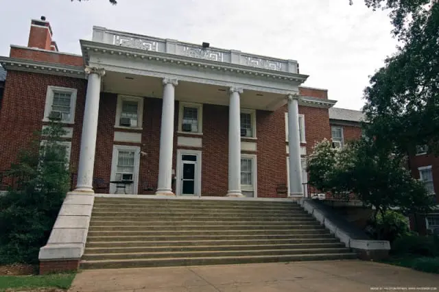 Brick building with a grand staircase leading to a portico supported by large white columns. A decorative balustrade tops the building under a cloudy sky.